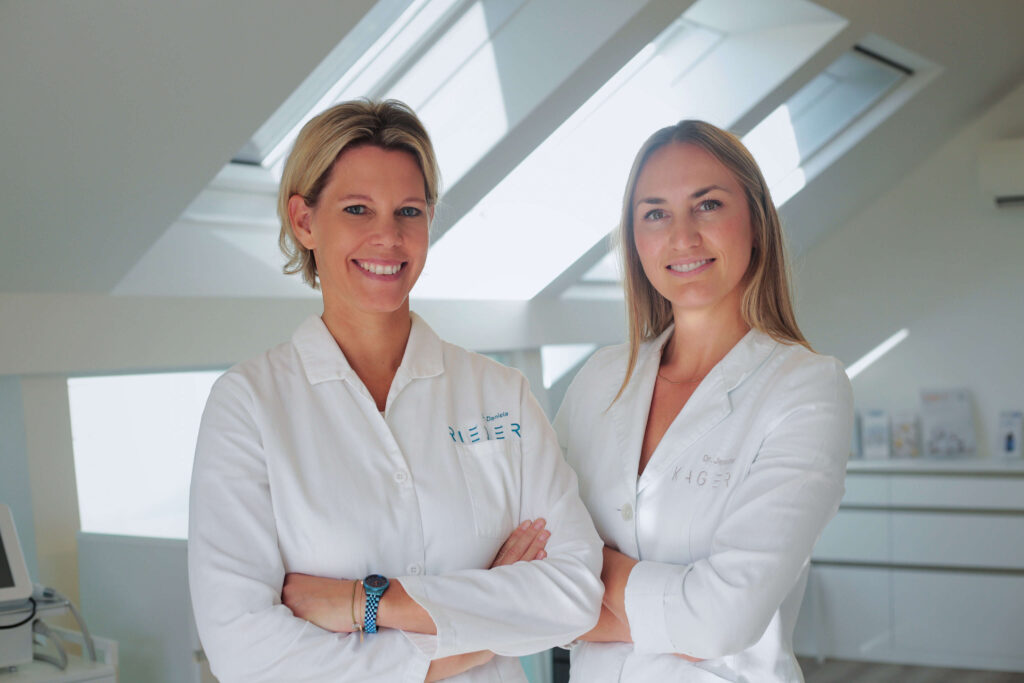 Dr. Daniela Rieder und Dr. Jennifer Kager, Spezialistinnen für ästhetische Medizin und Chirurgie, lächelnd in ihrer Praxis in weißen Kitteln.